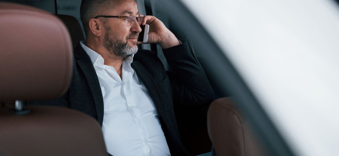 Calm and good mood. Having business call while sitting at the back of modern luxury car. Senior man in glasses and official wear.