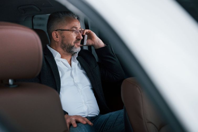 Calm and good mood. Having business call while sitting at the back of modern luxury car. Senior man in glasses and official wear